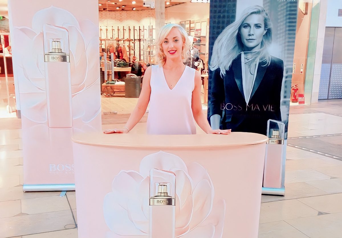 A promotional staff member standing behind a pale pink Boss Ma Vie sampling counter in a shopping centre