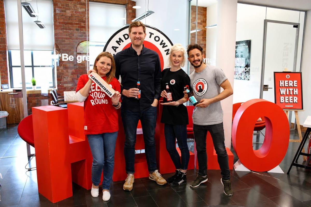 Four Camden Town Brewery team members holding beers beside giant red HELLO letters in an office