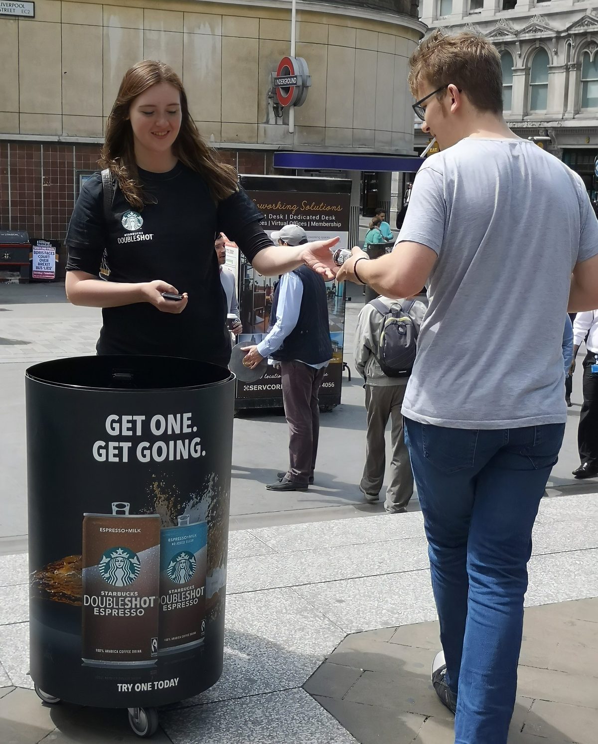 Starbucks sampling staff member handing a Doubleshot Espresso can to a commuter beside a branded Get One Get Going sampling bin
