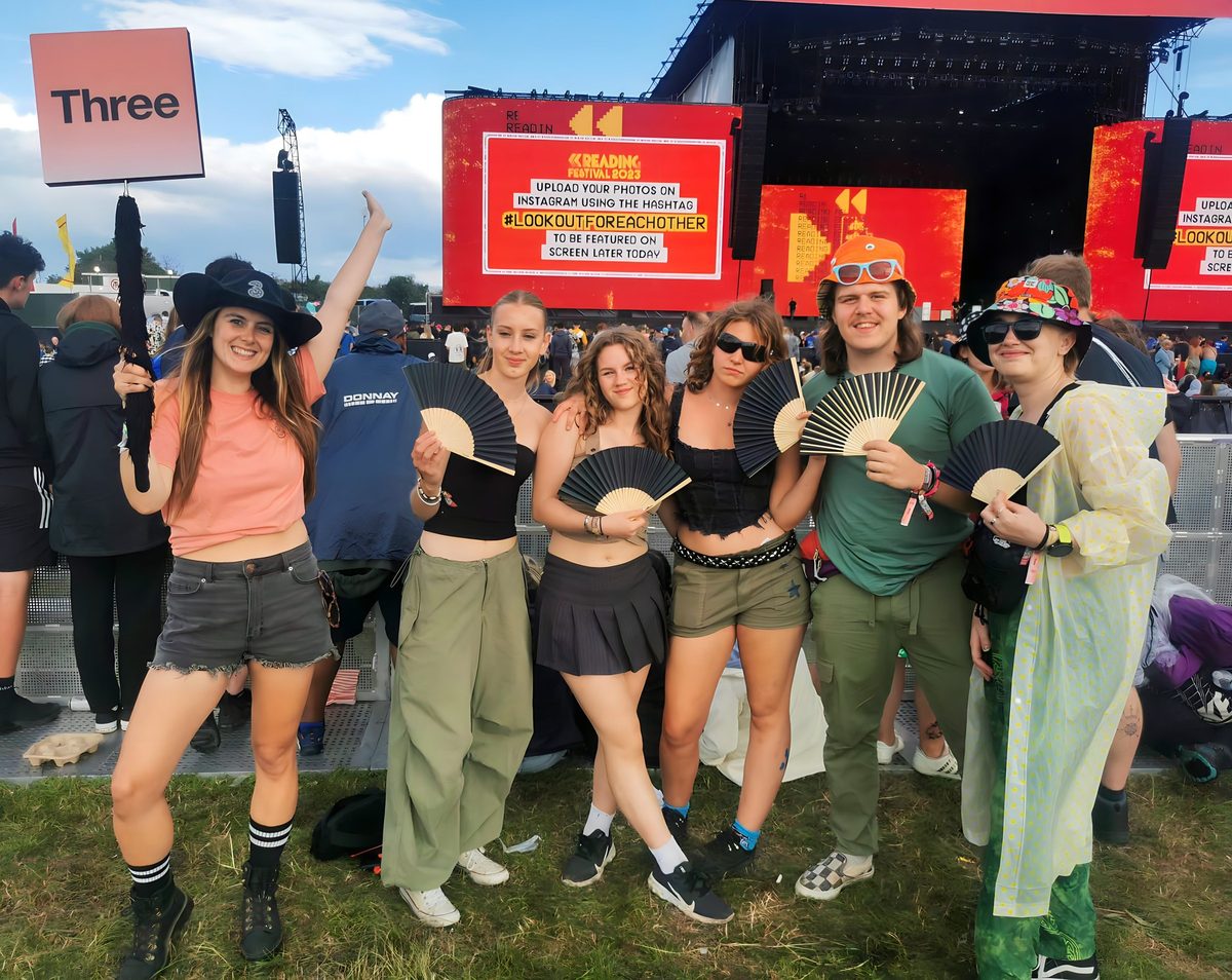 A large group of festival-goers with fans in front of the Three platform and main stage screen