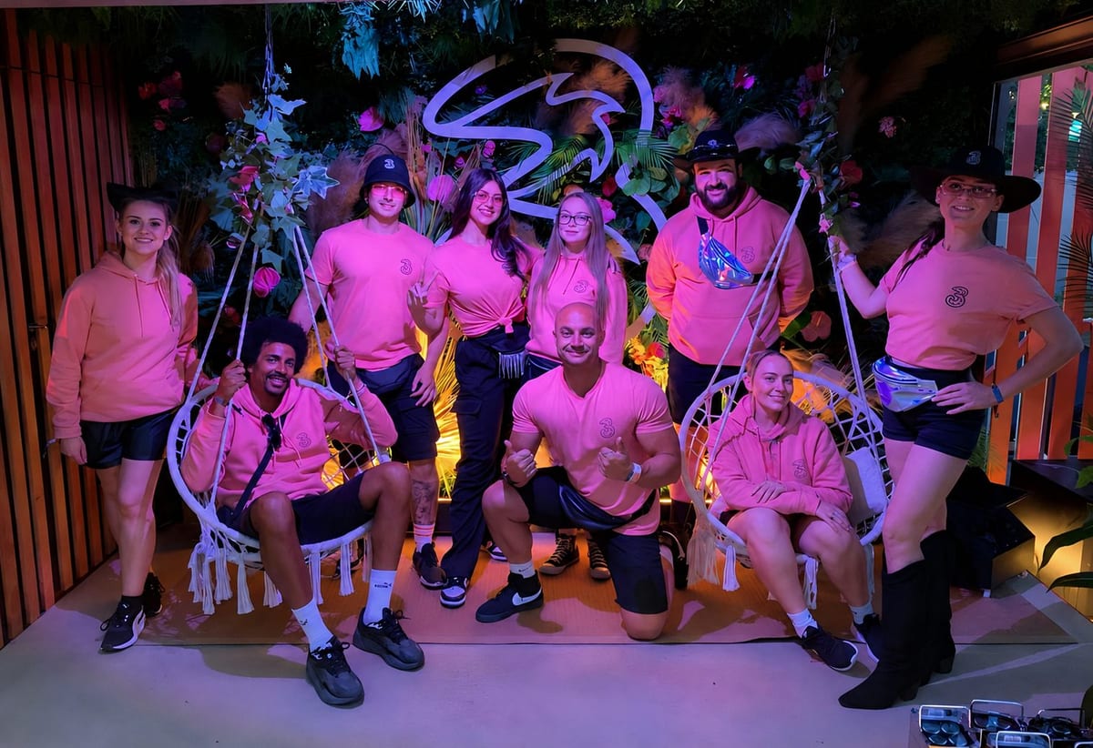 The Three UK festival team in pink tops posing on the lit VIP platform at night