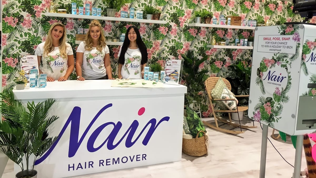 Three Nair brand ambassadors in white t-shirts behind the Nair Hair Remover counter on a tropical-themed stand