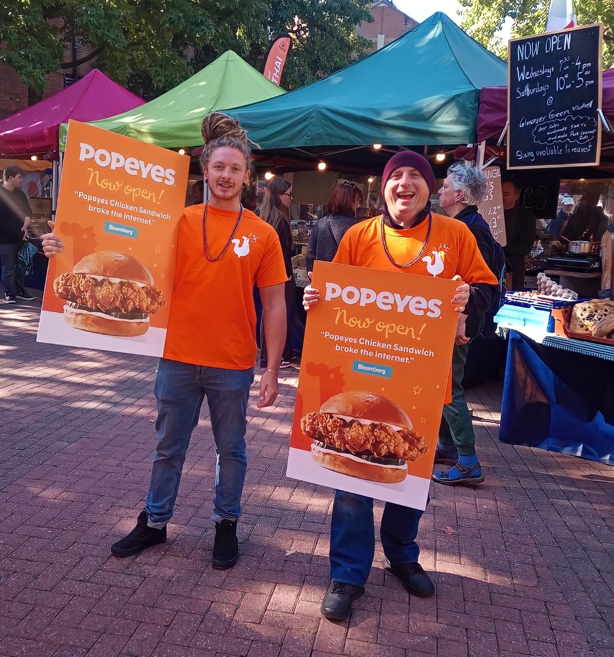Two promotional staff holding Popeyes posters at an outdoor market