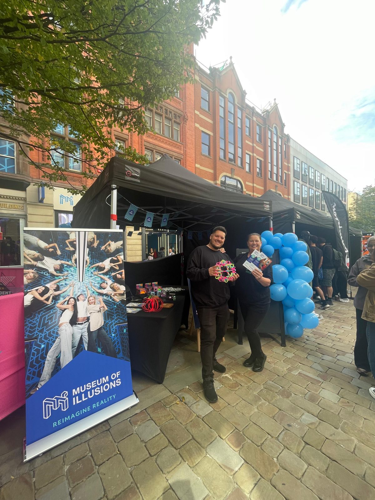 Two brand ambassadors holding Museum of Illusions leaflets by the branded gazebo on Market Street, Manchester